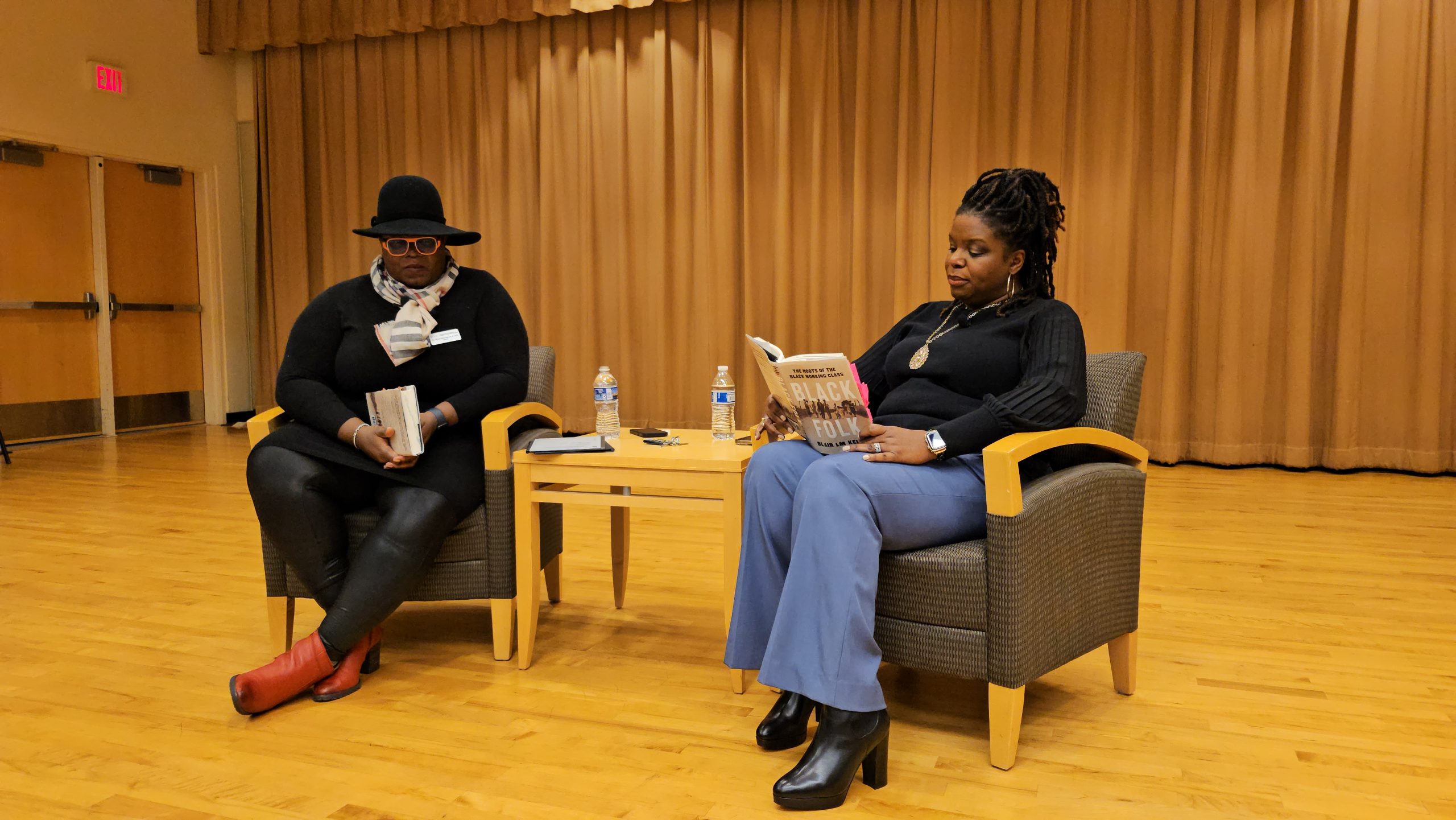 Photo of Kelley reading an excerpt from her book "Black Folk." Manigault-Bryant puts her head down as she listens.
