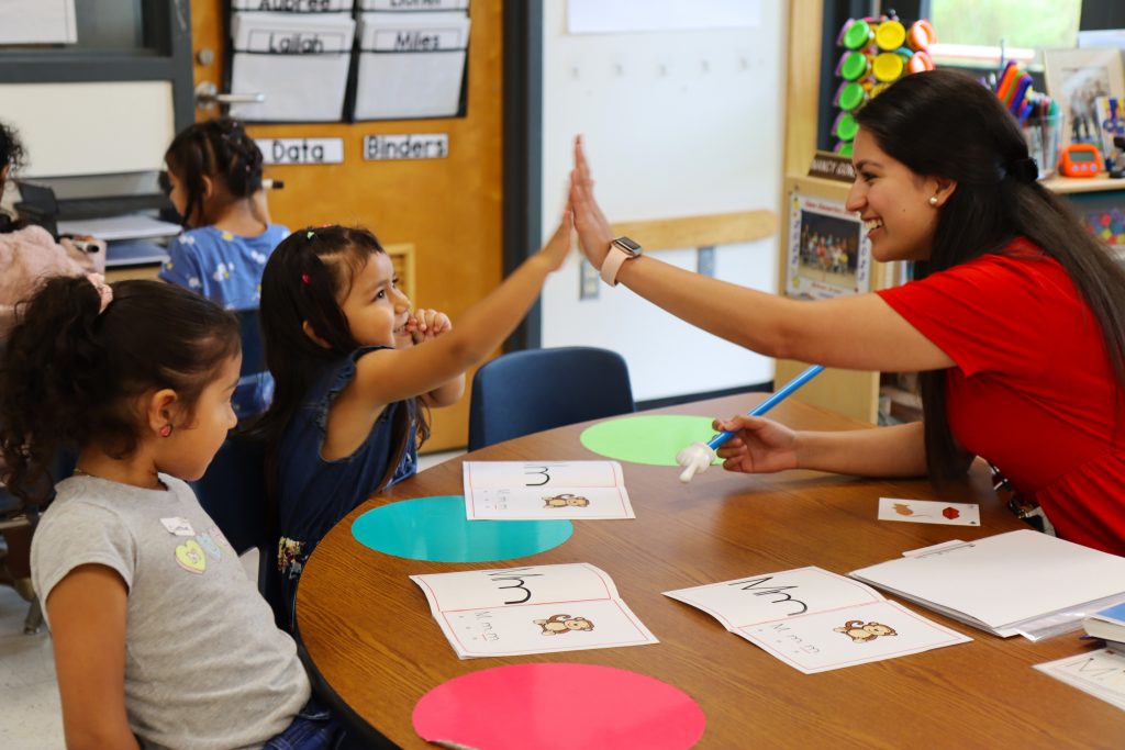 Photo of kindergarten teacher, Nancy Gonzalez high-fiving her student Angelyn Reyes Espino after getting a question about "M" sounds correct. Gonzalez's other student, Cinthia Argueta, looks at the high-five.