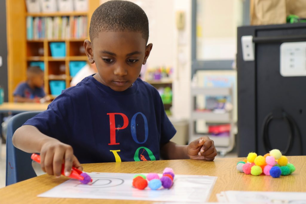 Close up of Legend Moody, kindergarten student of Nancy Gonzalez's using small solid colored pom-poms to fill in the letter "D" on a piece of paper as a learning exercise.