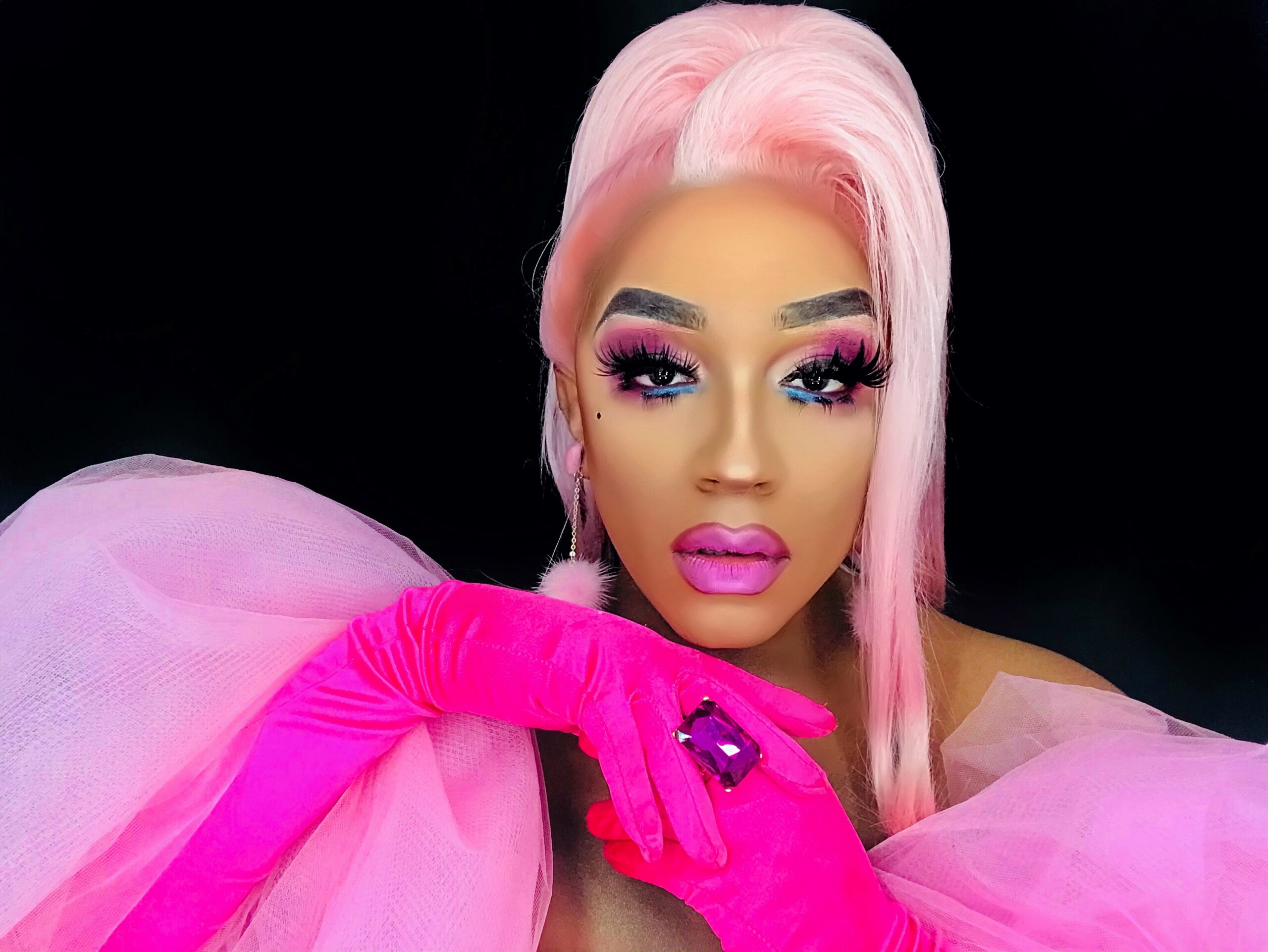 Naomi Dix, a drag queen, looks at the camera with her fingers crossed. She has pink hair, colorful pink-and-blue makeup, and is wearing a pink dress with hot pink gloves and a large ring.