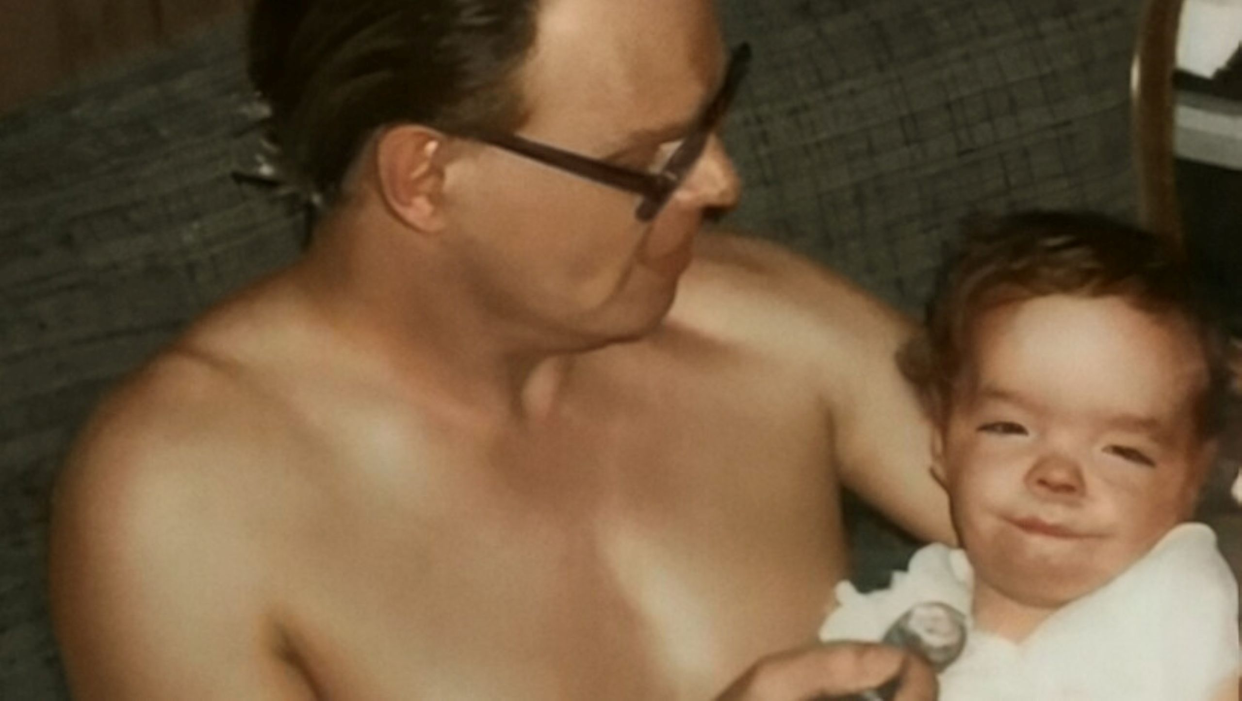 A shirtless white man with short dark-brown hair and black glasses holds a spoonful of food to a baby in his arms. The baby has curly brown hair and is wearing a white shirt and a diaper.